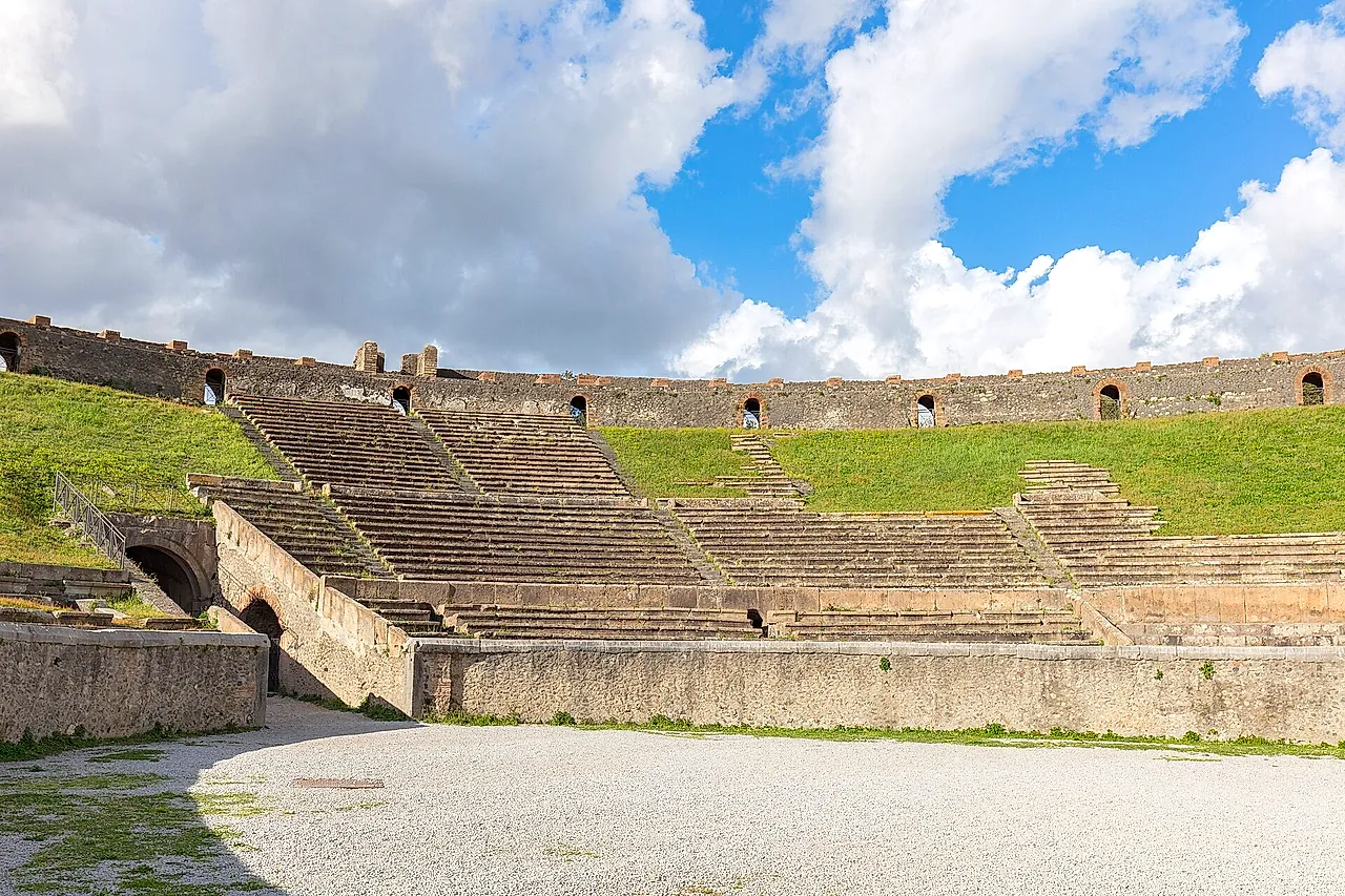 The arena and grass-covered seating of the Pompeii amphitheatre under a cloudy blue sky
