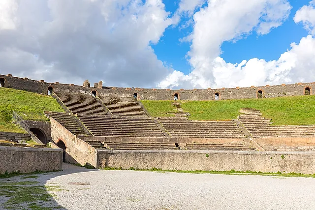 The arena and grass-covered seating of the Pompeii amphitheatre under a cloudy blue sky